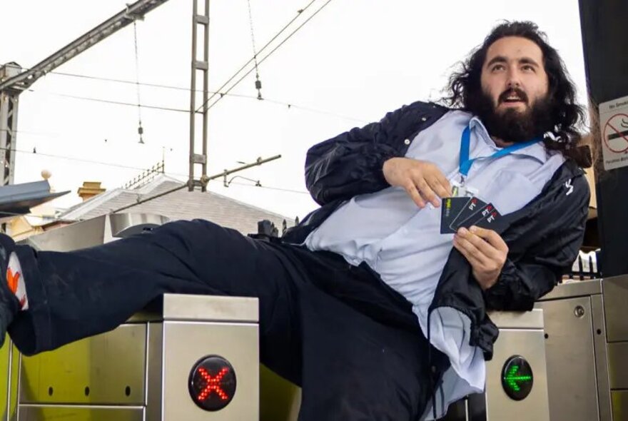 A bearded man dressed as a ticket inspection lounging over the gates of a station and holding Myki cards.
