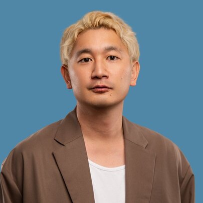 Comedian Takashi Wakasugi, wearing a brown jacket over a white t-shirt, he has a neutral facial expression, blond hair and stands against a blue background.