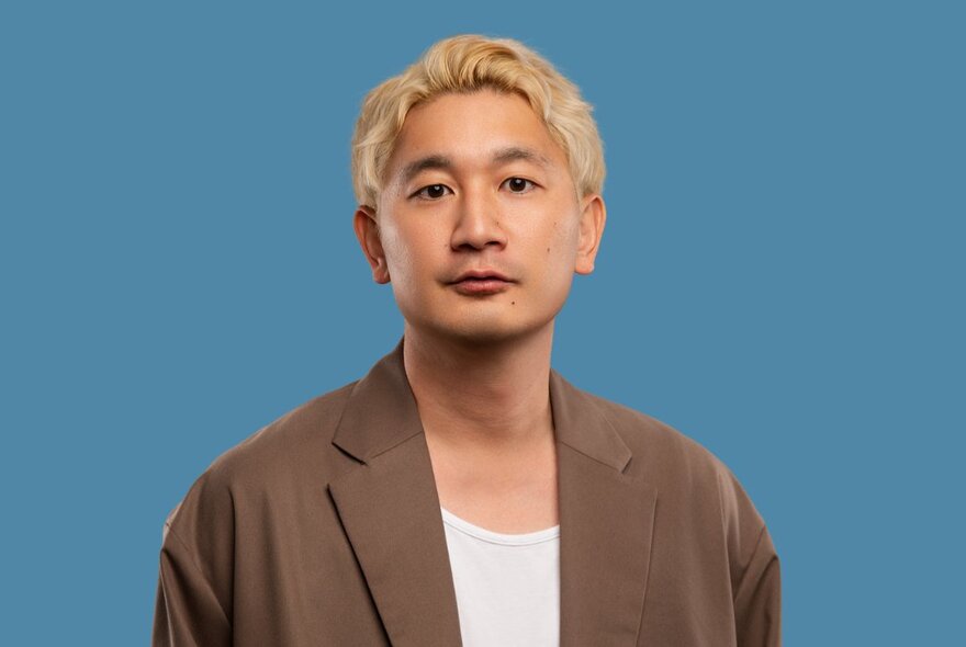 Comedian Takashi Wakasugi, wearing a brown jacket over a white t-shirt, he has a neutral facial expression, blond hair and stands against a blue background.