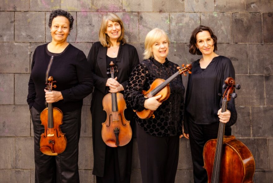 The four members of the Flinders Quartet, smiling while standing against a wall and holding their musical instruments.