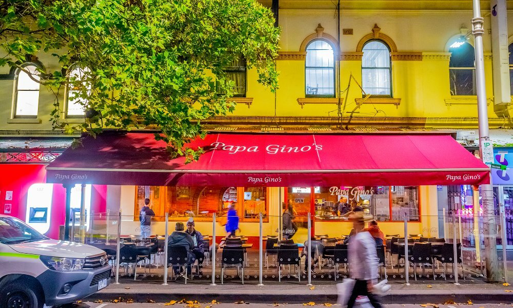 People walking past a restaurant with outdoor seating and a large red awning at night.