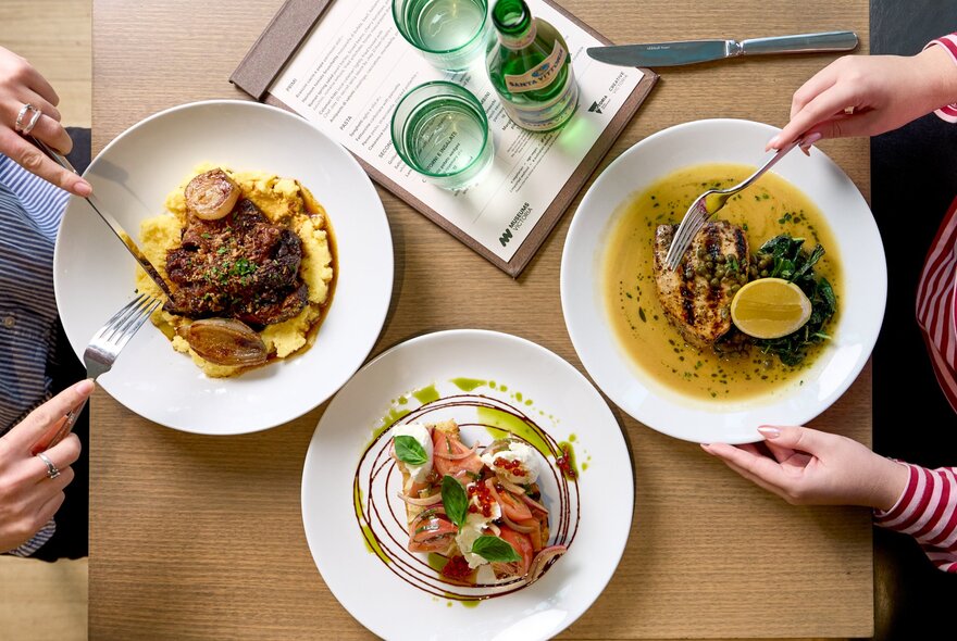 Looking down on a table with hands holding cutlery over two main courses with a side salad and tray of mineral water. 