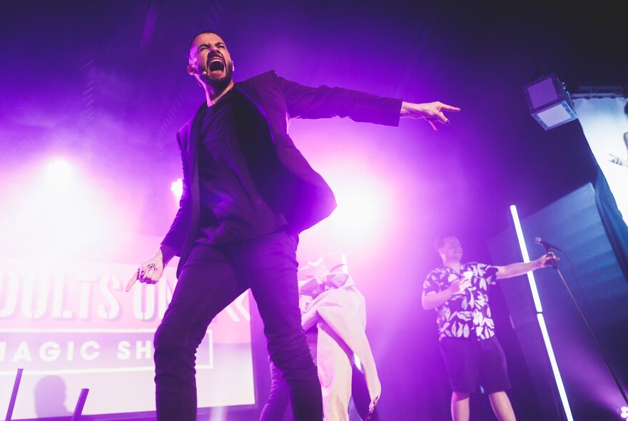 A male performer in a black suit, with his mouth open, stands on a stage under bright purple lights, pointing his right hand toward the audience, with people on the stage behind him.