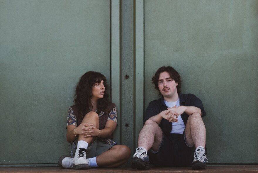 A man and woman sitting on the ground, with their backs leaning up against a wall, both in a relaxed pose.