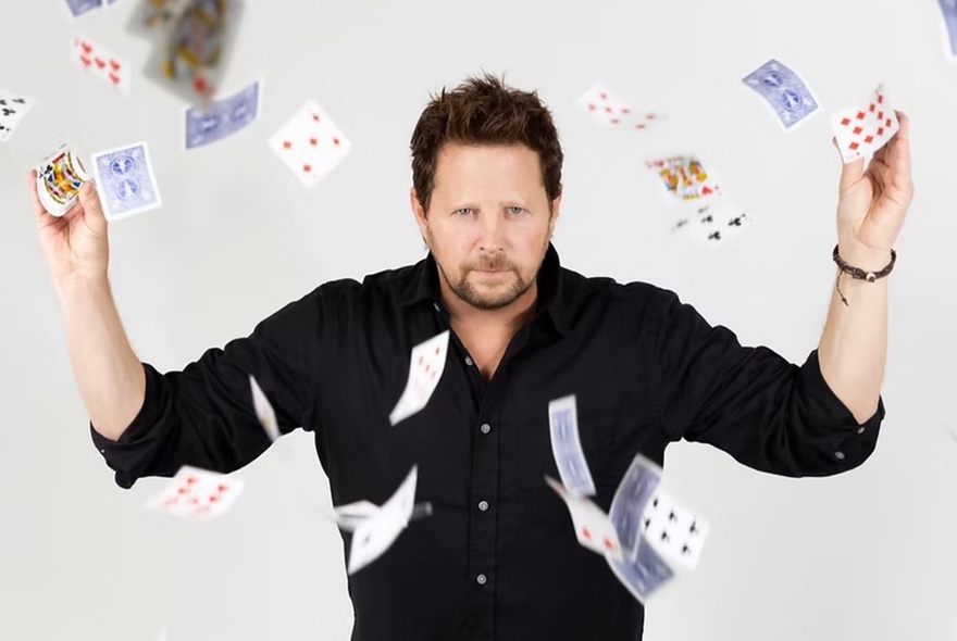 A man staring straight ahead with his hands up and flipping playing cards so that they rain down around him.