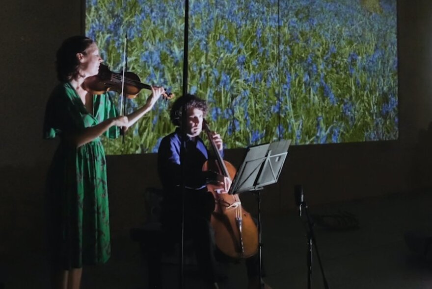 A musical performance featuring a cellist and a violinist playing on a stage with a scenic landscape projected on a large screen behind them.
