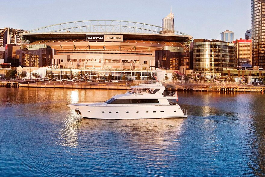 A white motor boat in the Docklands Harbour, with Marvel Stadium in the background. 