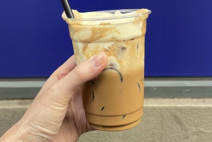 A hand holding a plastic cup of iced Vietnamese Salt Coffee with a black strawer, against a blue and grey background.