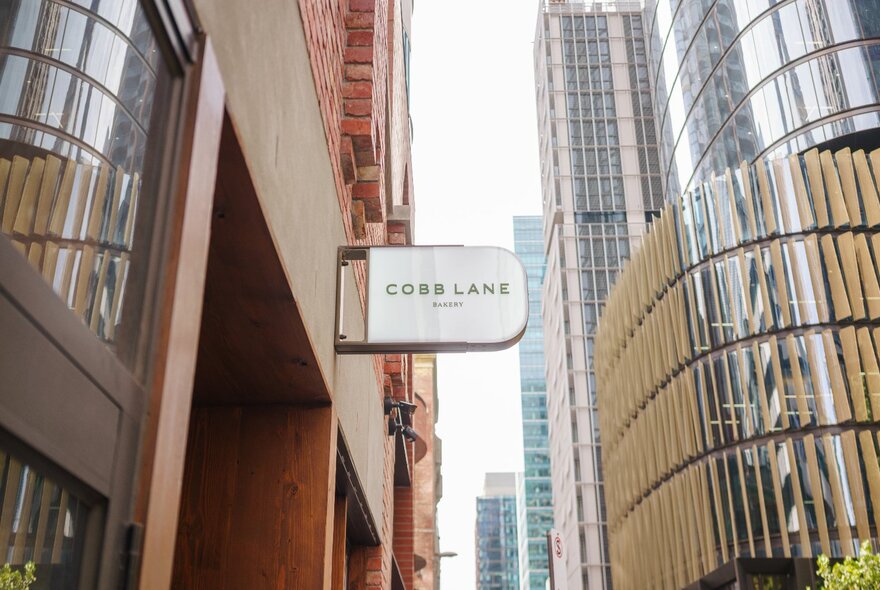 The sign for Cobb Lane bakery in a city street with skyscrapers all around. 