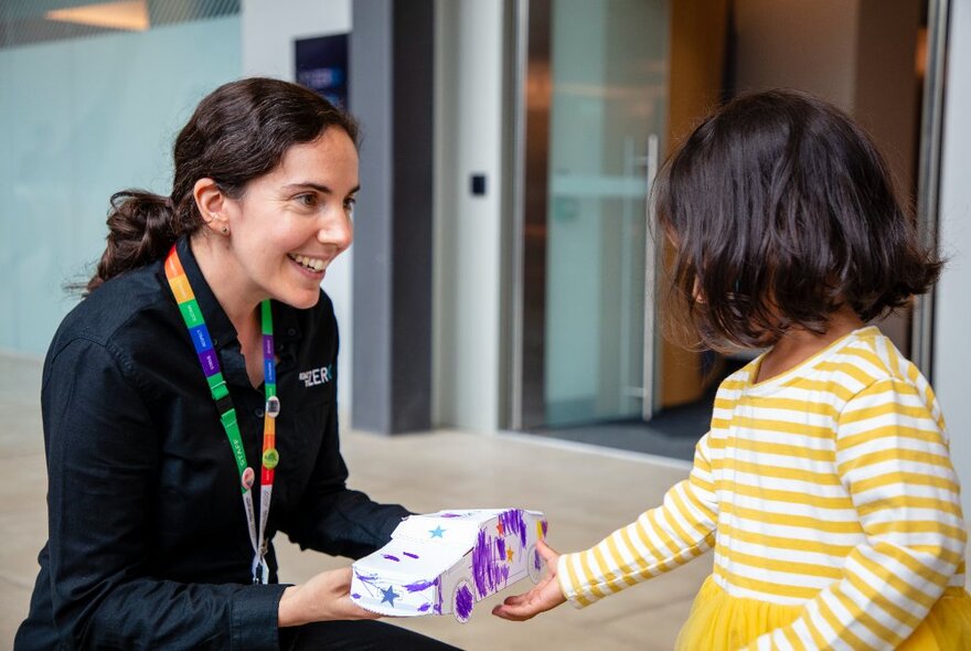 A young child accepting a decorated cardboard car from a Museum Victoria staff member dressed all in black.