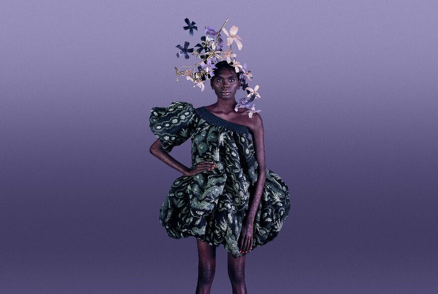 An Indigenous model in a one-shouldered puffy dress with a large headpiece of shiny flowers. 