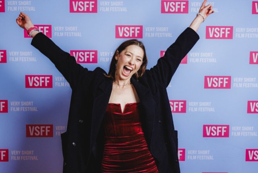 Woman standing in front of signage with arms raised, smiling widely.