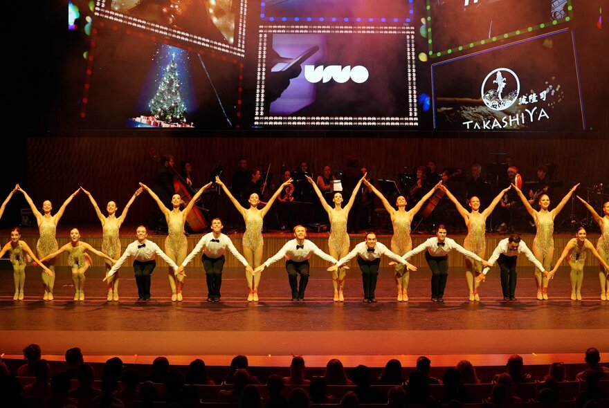 Rows of dancers performing with arms oustretched, on stage.