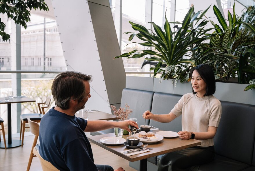 A couple eating together in a bright, modern restaurant setting with large windows and indoor plants.