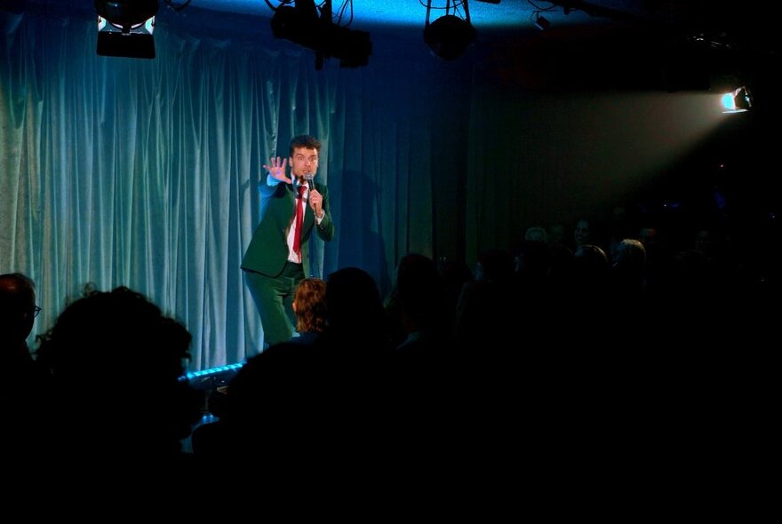 Comedian Oliver Coleman delivering a stand up routine on stage with a blue curtain behind him.