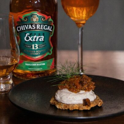 A whisky cocktail in a tall champagne glass alongside a nip of whisky in a stemless cognac glass, with a cracker with toppings on a black plate, a bottle of whisky in the background.