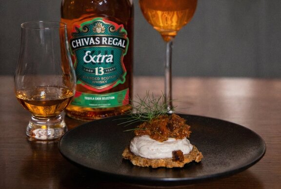 A whisky cocktail in a tall champagne glass alongside a nip of whisky in a stemless cognac glass, with a cracker with toppings on a black plate, a bottle of whisky in the background.