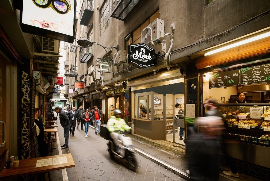 Centre Place laneway showing the brightly lit shop exterior of Mork Chocolates, with hanging signage, and pedestrians walking past.