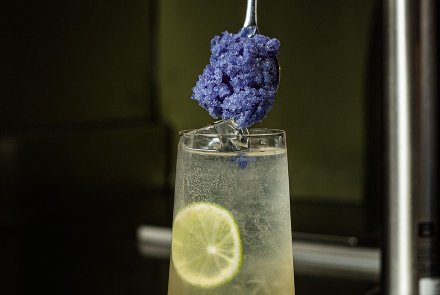 A lime and soda cocktail with a spoon full of crushed frozen blueberries about to be dipped into it.