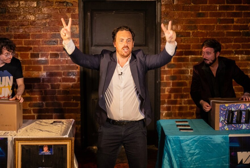 Host on a stage during a performance of the live comedy game show IKEA Wars, with two tables behind him, each with a person about to assemble furniture from a flatpack box.