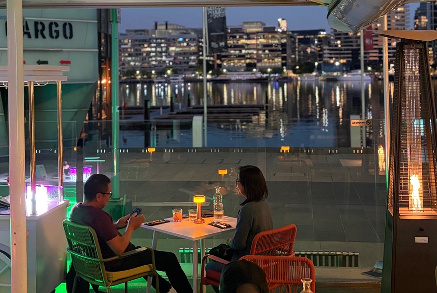 Diners seated outside at dusk overlooking Docklands harbour with city buildings reflected in the water.