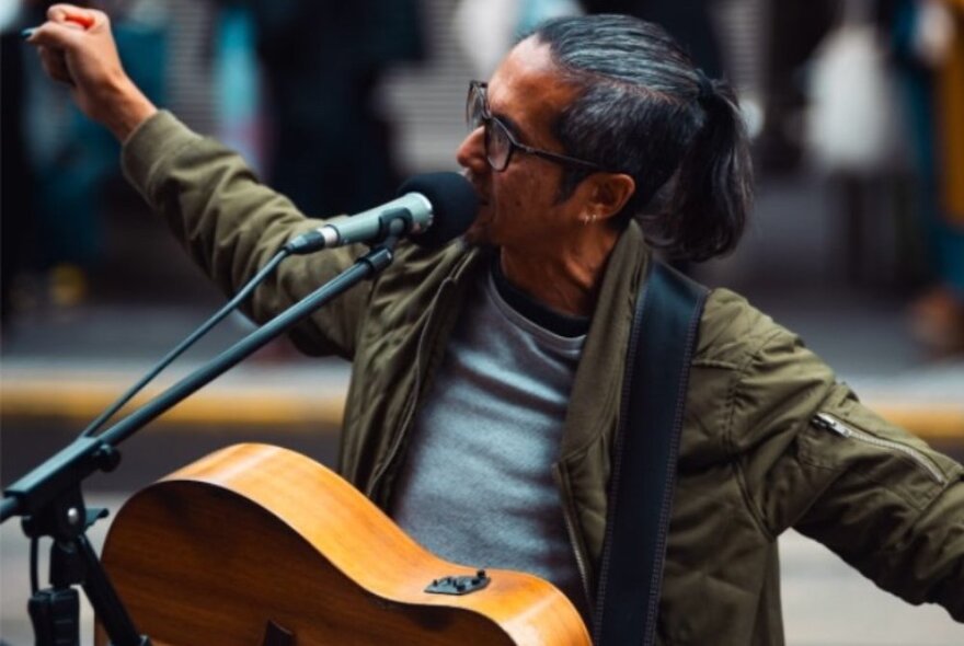 A busker performing in a street with a guitar around his neck and arms outstretched while singing. 
