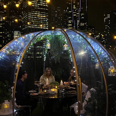 A group of four patrons dining in a heated glass dome on a rooftop with Melbourne's city skyline at night behind them. 