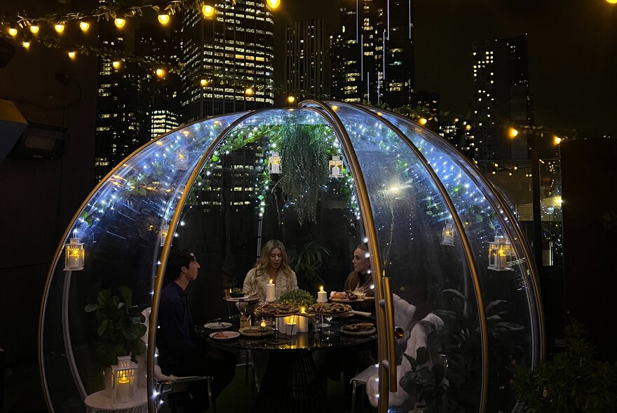 A group of four patrons dining in a heated glass dome on a rooftop with Melbourne's city skyline at night behind them. 