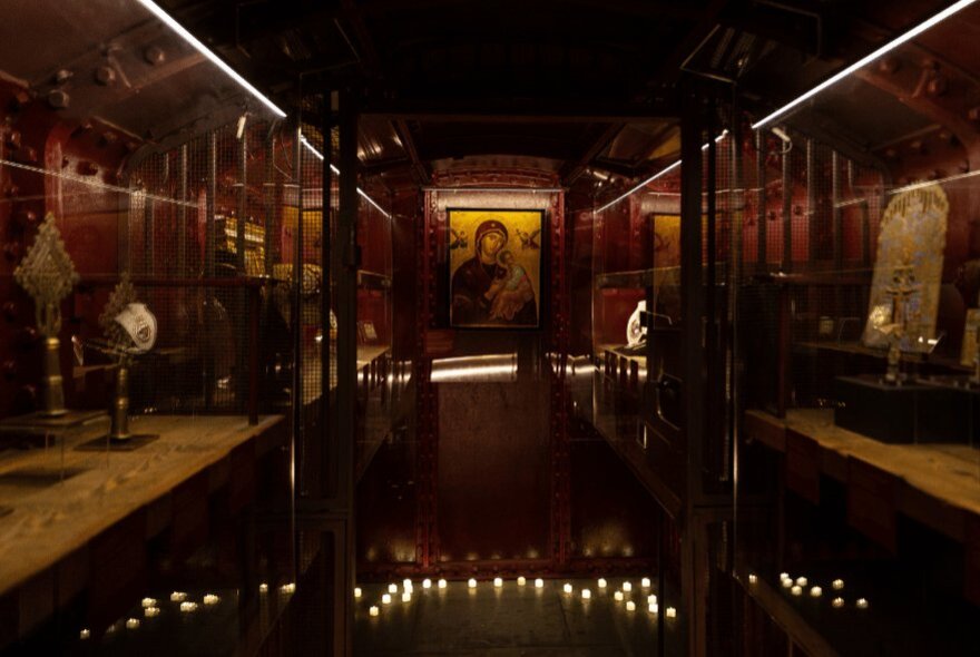 A dimly lit museum gallery featuring rows of wooden display cases along the sides and small candles or lights illuminating the floor at the bottom of the cases.