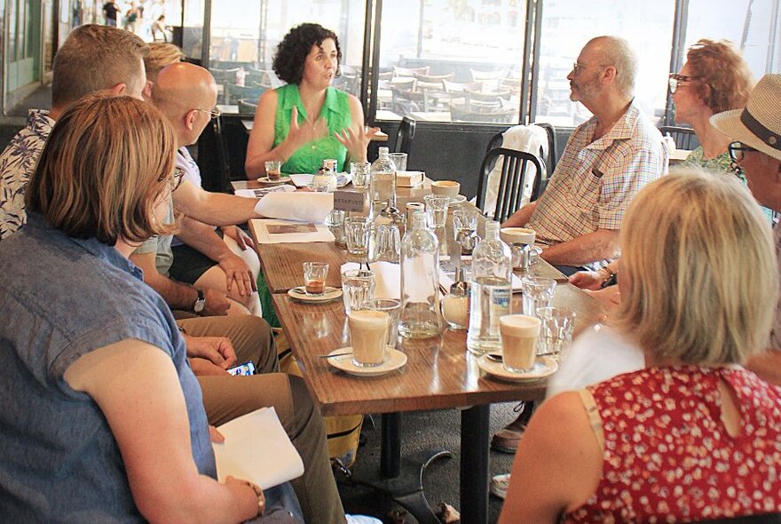 A tour group listening to a speaker, seated at a dining table with drinks.