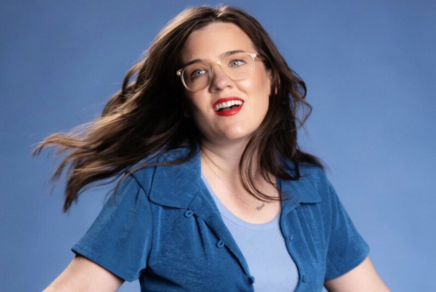 Comedian Rose Callaghan, with her straight brown hair flowing in the air behind her, head tilted to one side, smiling slightly, wearing a blue top and glasses.