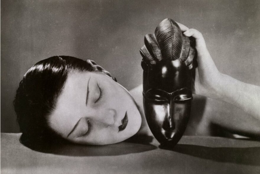 A black and white example of a Surrealist artwork, featuring a woman resting her head on a table whilst holding a traditional wooden mask.