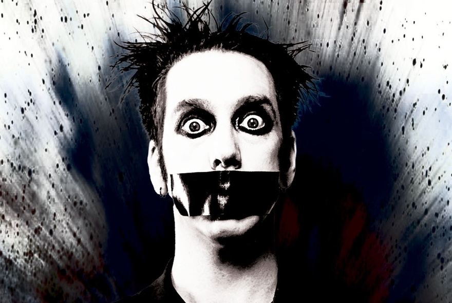 The face of a man wearing thick black eye make-up and black tape across his mouth, eyes widened in surprise; a black starburst in the background.