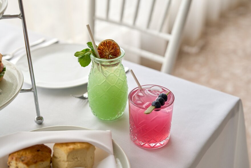 Two vibrant beverages, a green drink garnished with an orange slice and mint, and a pink drink topped with berries.