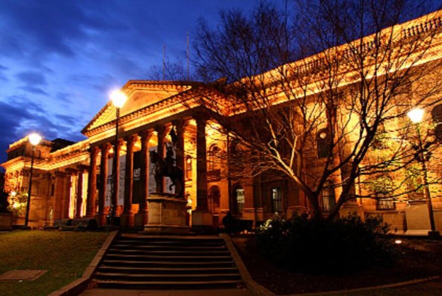 State Library Victoria exterior lit at night with bare tree, lamp lighting and cloudy sky.