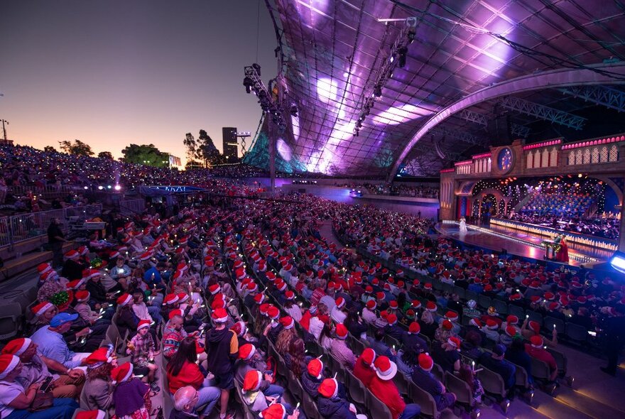 Seated crowds in front of illuminated Myer Music Bowl stage, audience wearing Santa hats at sunset.