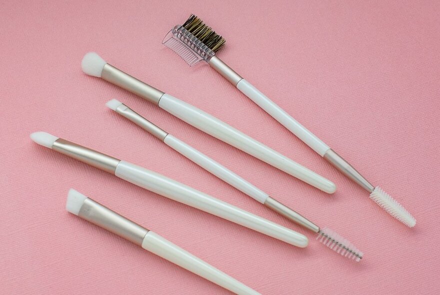 Five makeup brushes with white handles against a pink background. 