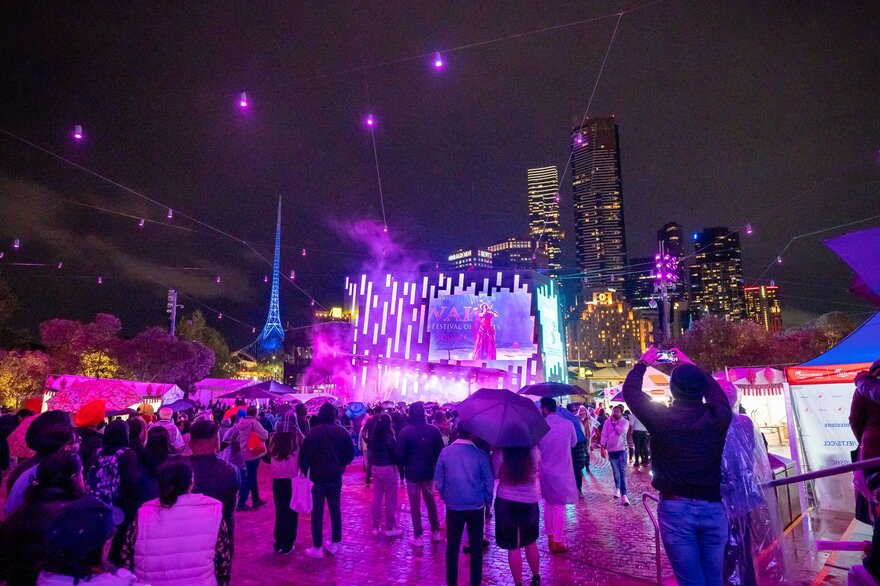 A crowd of people with umbrellas watching a purple-lit stage outdoors.