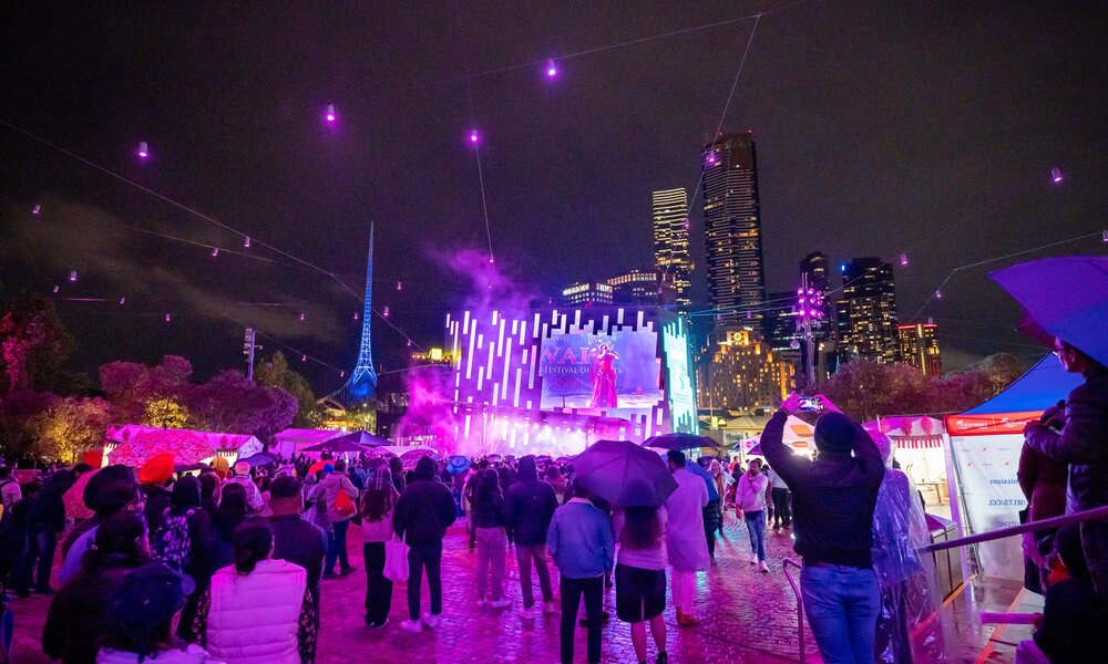 A crowd of people with umbrellas watching a purple-lit stage outdoors.