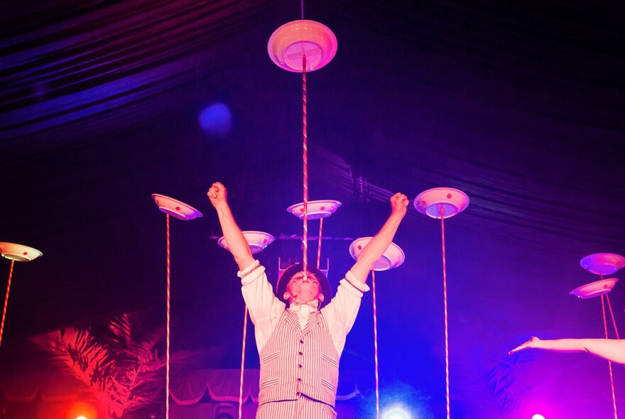 A circus perfomer balancing a plate on a pole in his mouth, other plates on poles in the background. 
