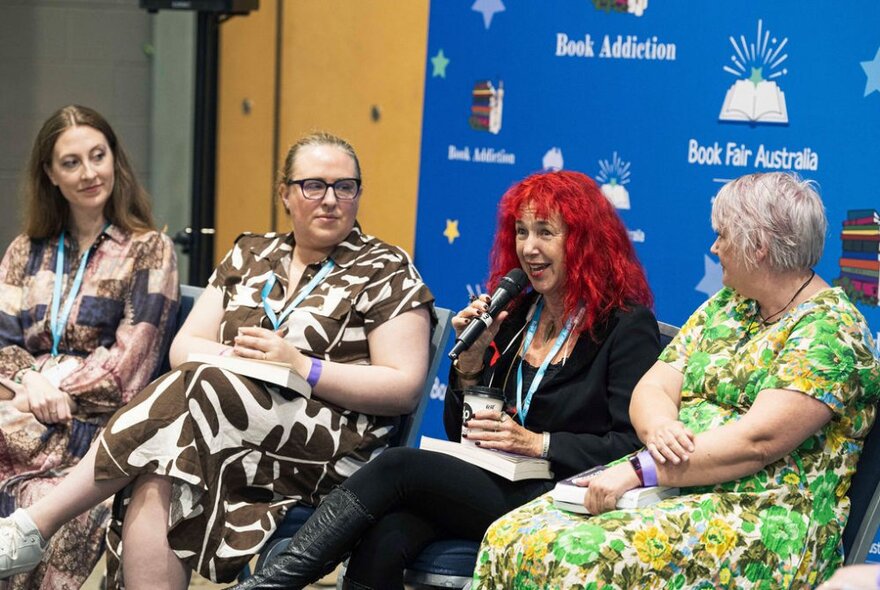 A panel of four women seated in front of a blue Book Fair Australia screen, each with a book in hand and one speaking into a microphone.