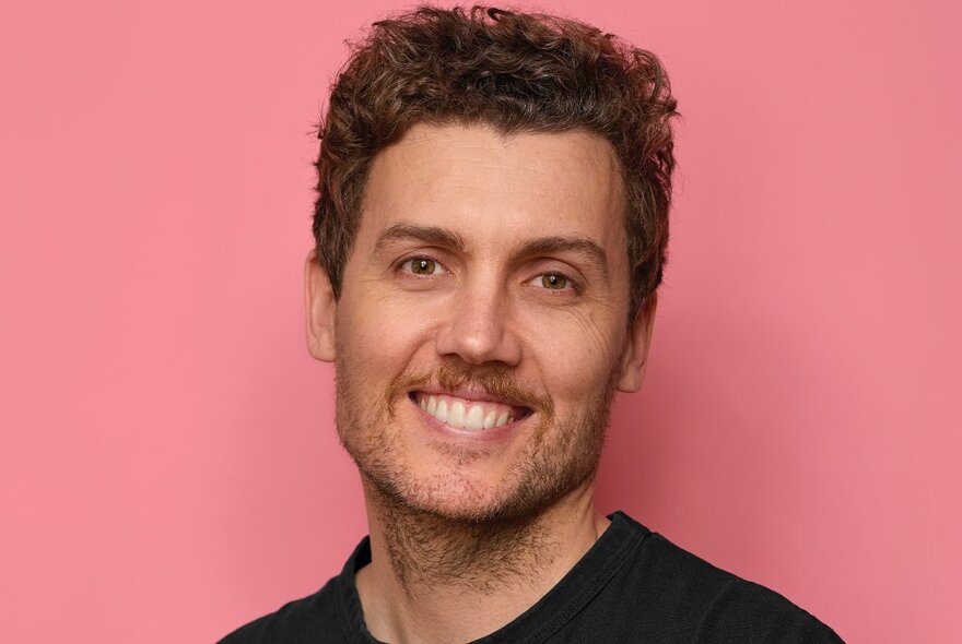 A man with short brown hair and a moustache, smiling at the camera and showing his teeth, posed against a pink background.