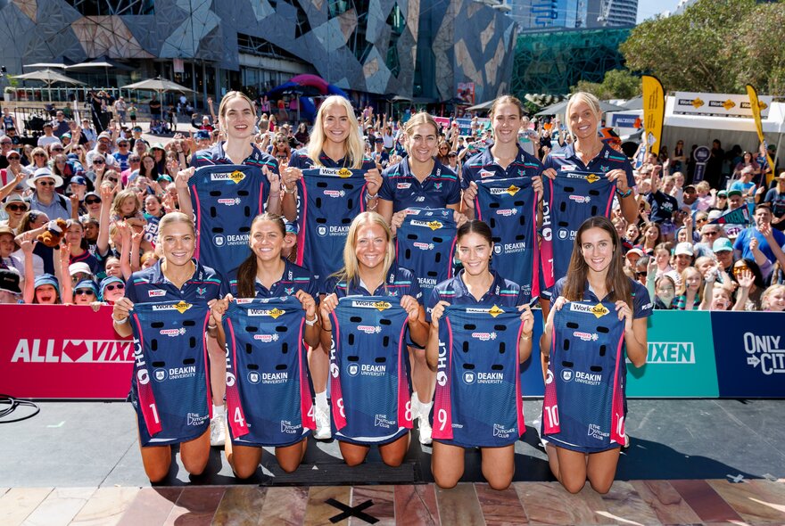 The Melbourne Vixens netball team posing for a group shot with an audience in Fed Square looking on. 