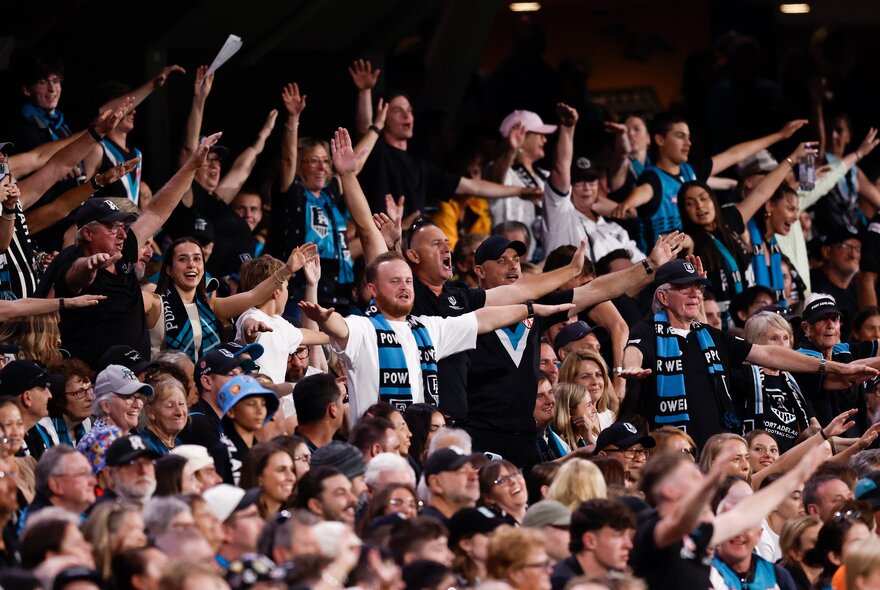 A crowd of AFL fans at a sports stadium, some of them standing with arms in the air, wearing Port Adelaide football club scarves and colours. 