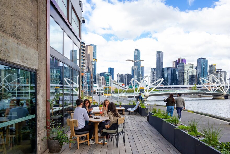 A group of friends are sitting outside at waterfront bar. There is a view of Melbourne's city skyline in the background.