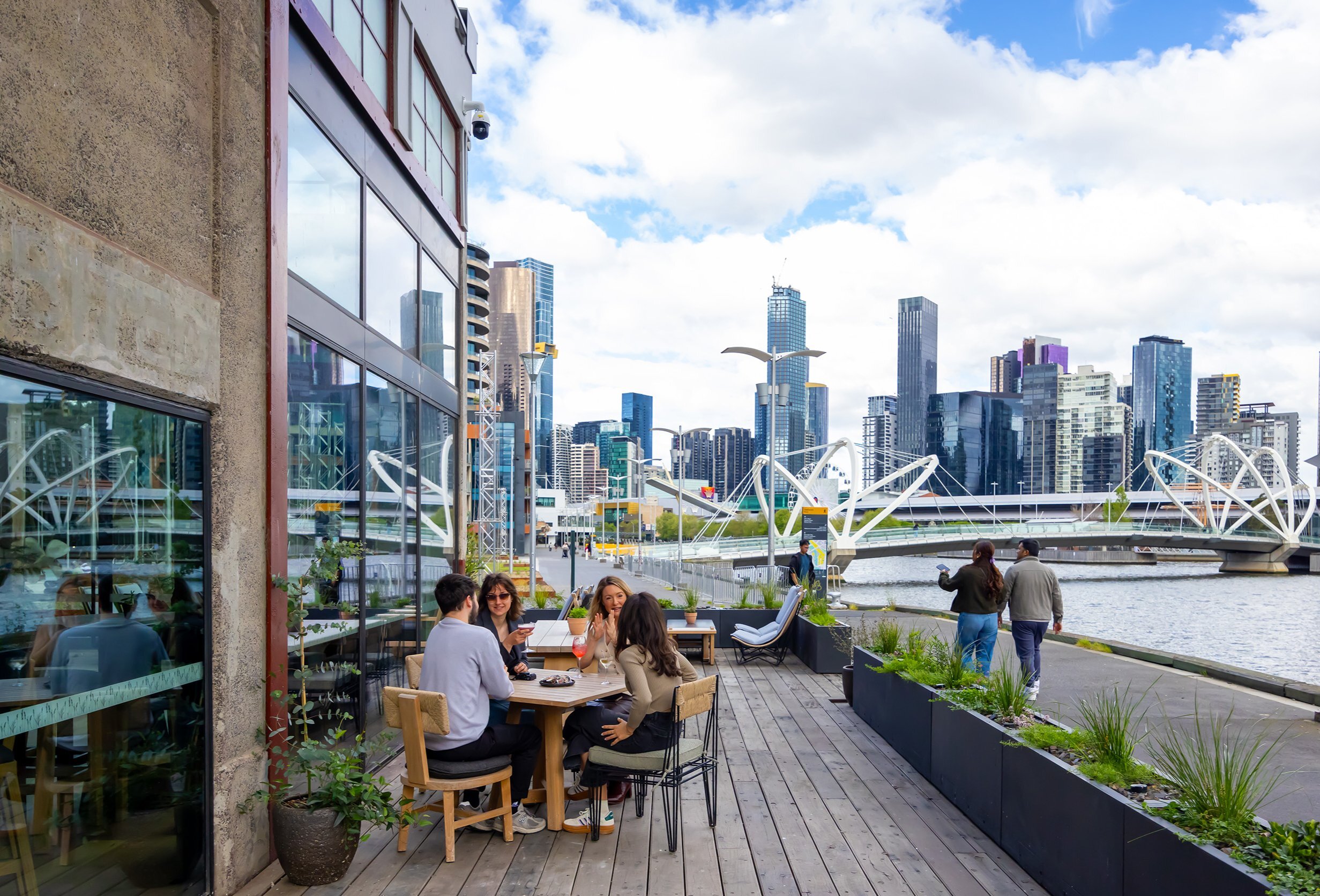 A group of friends are sitting outside at waterfront bar. There is a view of Melbourne's city skyline in the background. 