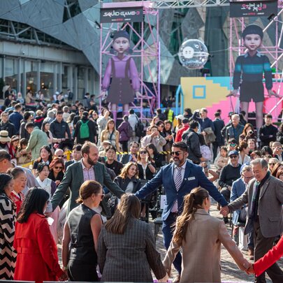 A group of people standing in a circle with their hands clasped and a large crowd of people behind them at Fed Square in Melbourne.