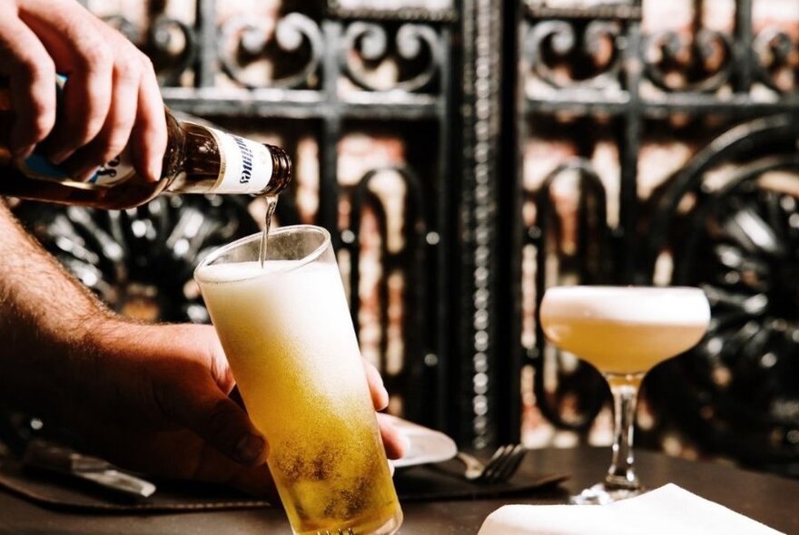 A bartender pouring a beer into a glass with a cocktail in the background.