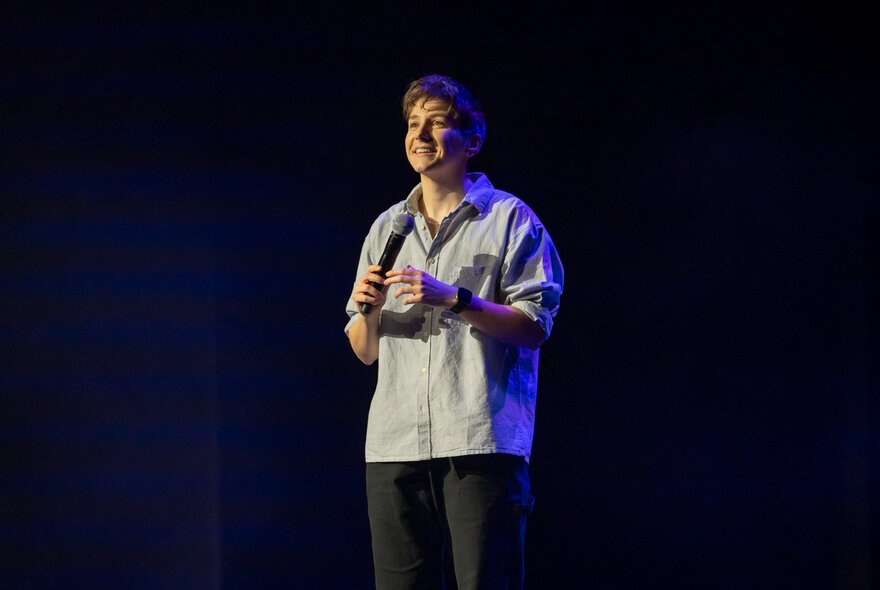 A young person on a dark stage, Sarah Keyworth, holding a microphone and smiling.