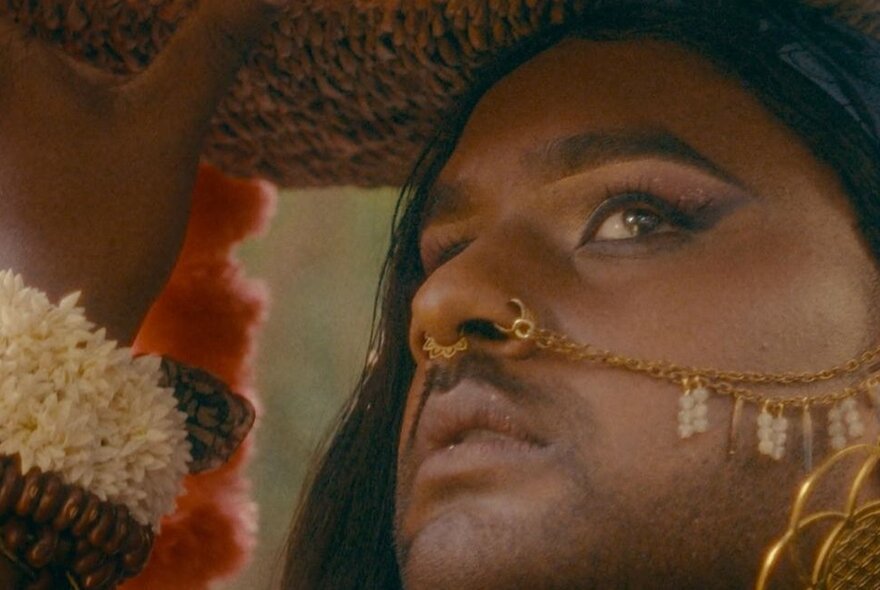 A person with a nose ring that attaches with a chain to to their (unseen) ear, looking cautiously upwards while holding a basket above their head. 